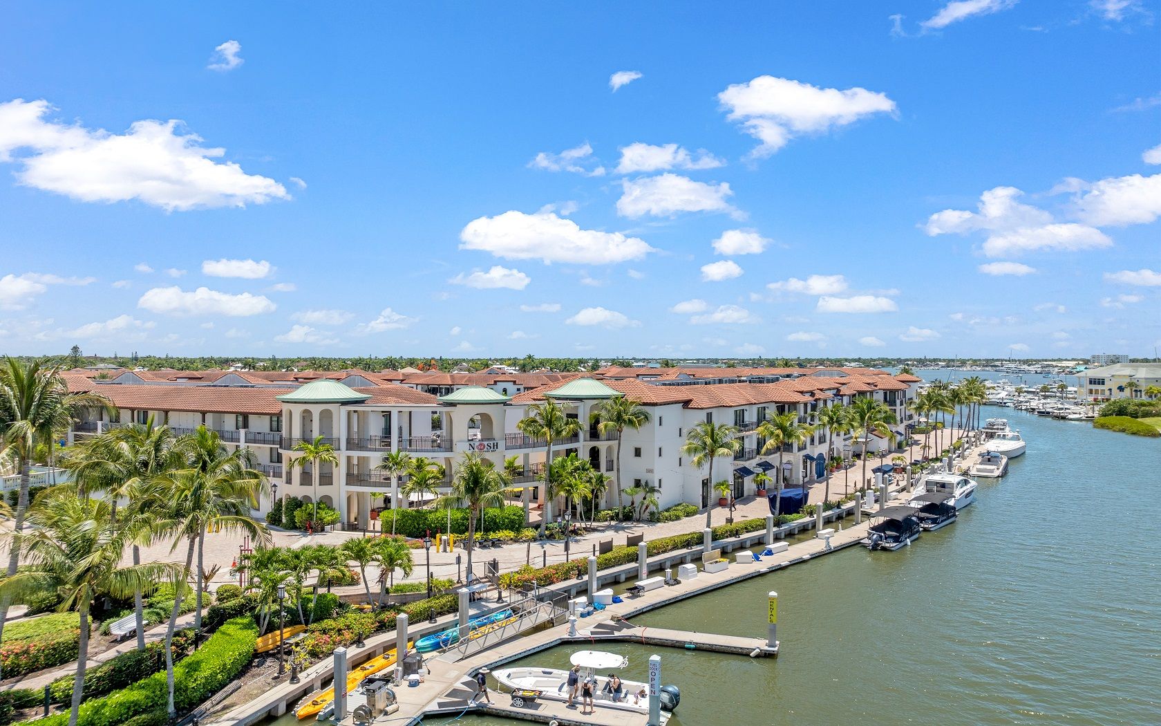 A sunny coastal marina with palm trees, white buildings, and boats docked along a canal by a blue sky&mdash;perfect day by the water.