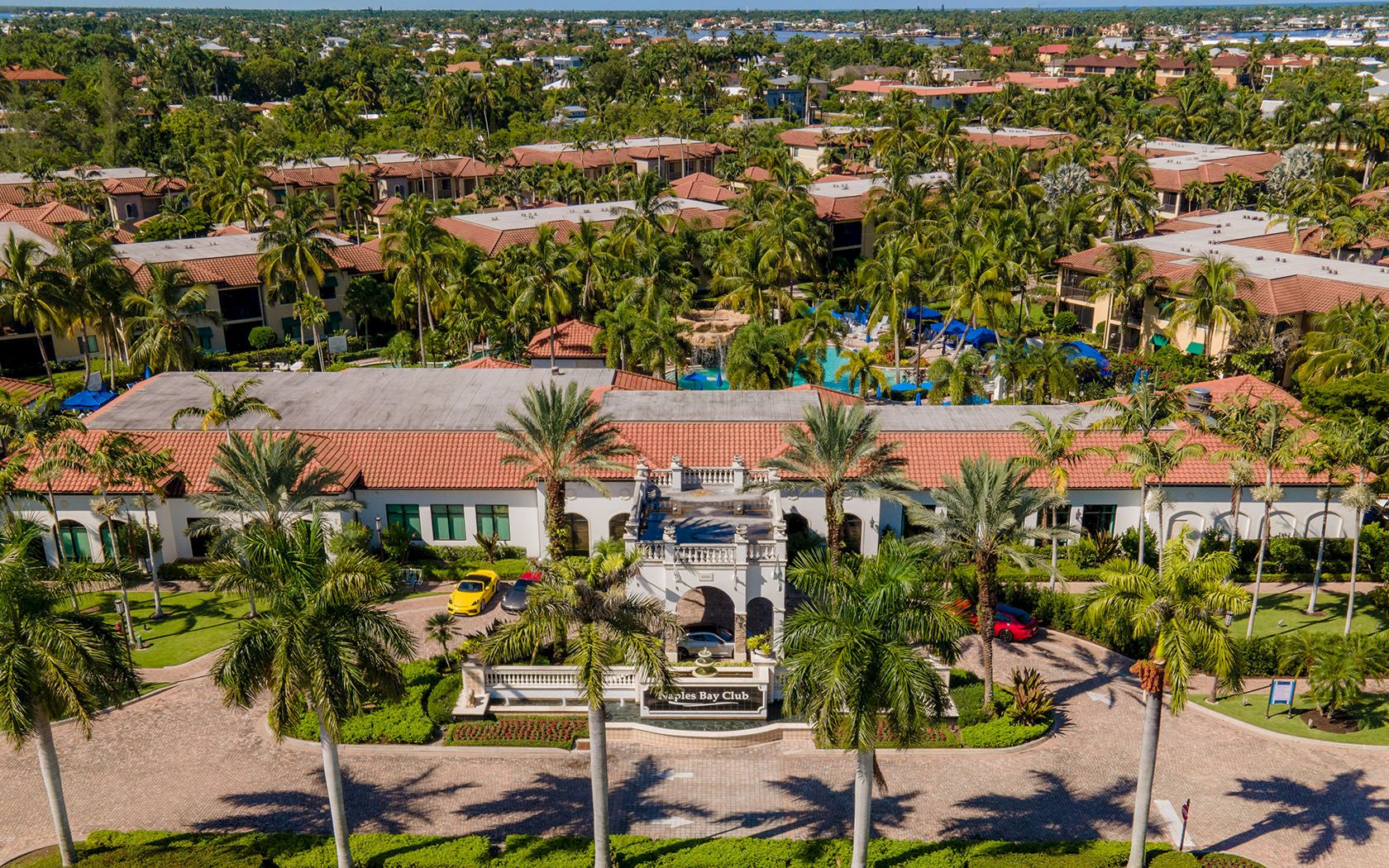 An aerial view of a resort with red-tiled roofs, palm trees, a central entry plaza, and a large pool area amid lush greenery, sunny and tropical.