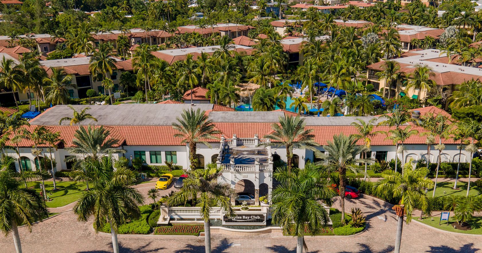 An aerial view of a resort with red-tiled roofs, palm trees, a central entry plaza, and a large pool area amid lush greenery, sunny and tropical.