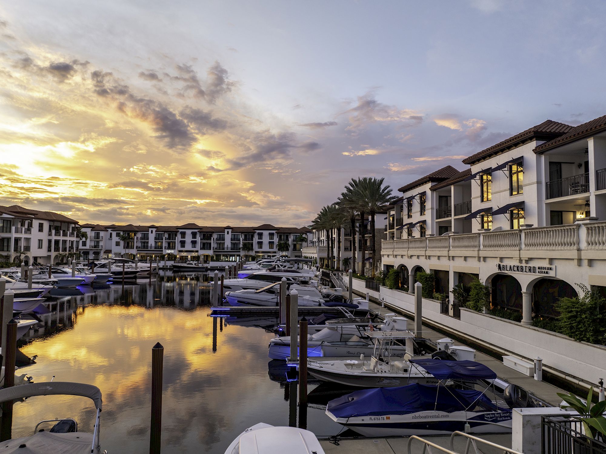 A luxury marina at sunset, with boats docked and white resort buildings lining the calm water.