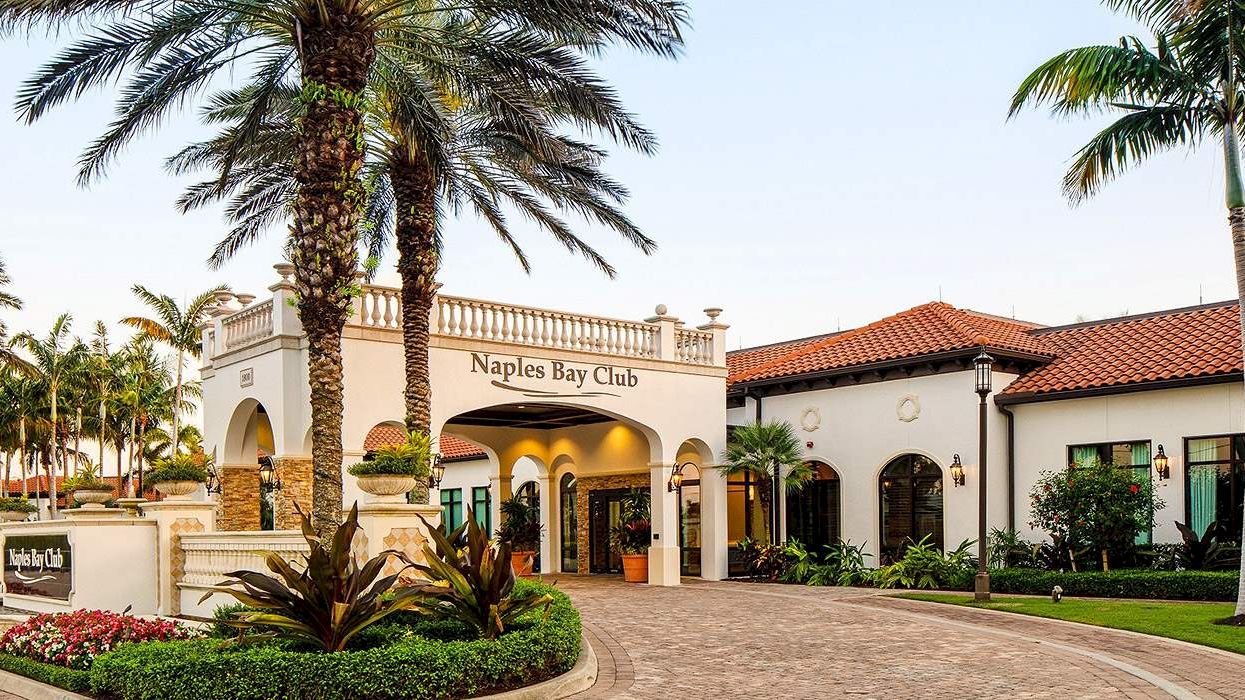A sunny resort entrance with palm trees, a Mediterranean-style building labeled &ldquo;Naples Bay Club,&rdquo; and a landscaped, curved brick driveway.