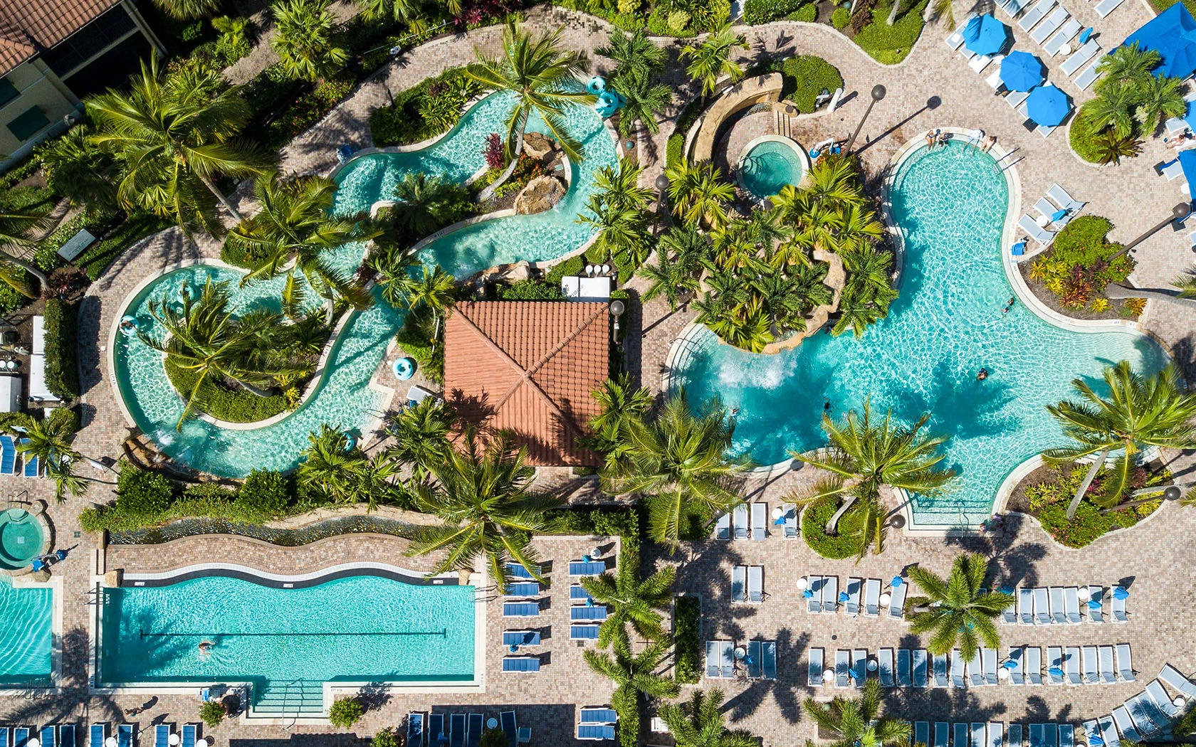 An aerial view of a resort pool area with winding pools, palm trees, sun loungers, and a central building, all surrounded by lush greenery.