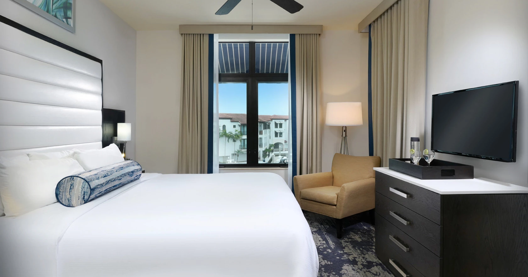Modern hotel room with a large bed, nightstand lamps, a patterned rug, a chair, floor-to-ceiling windows, and a wall-mounted TV.