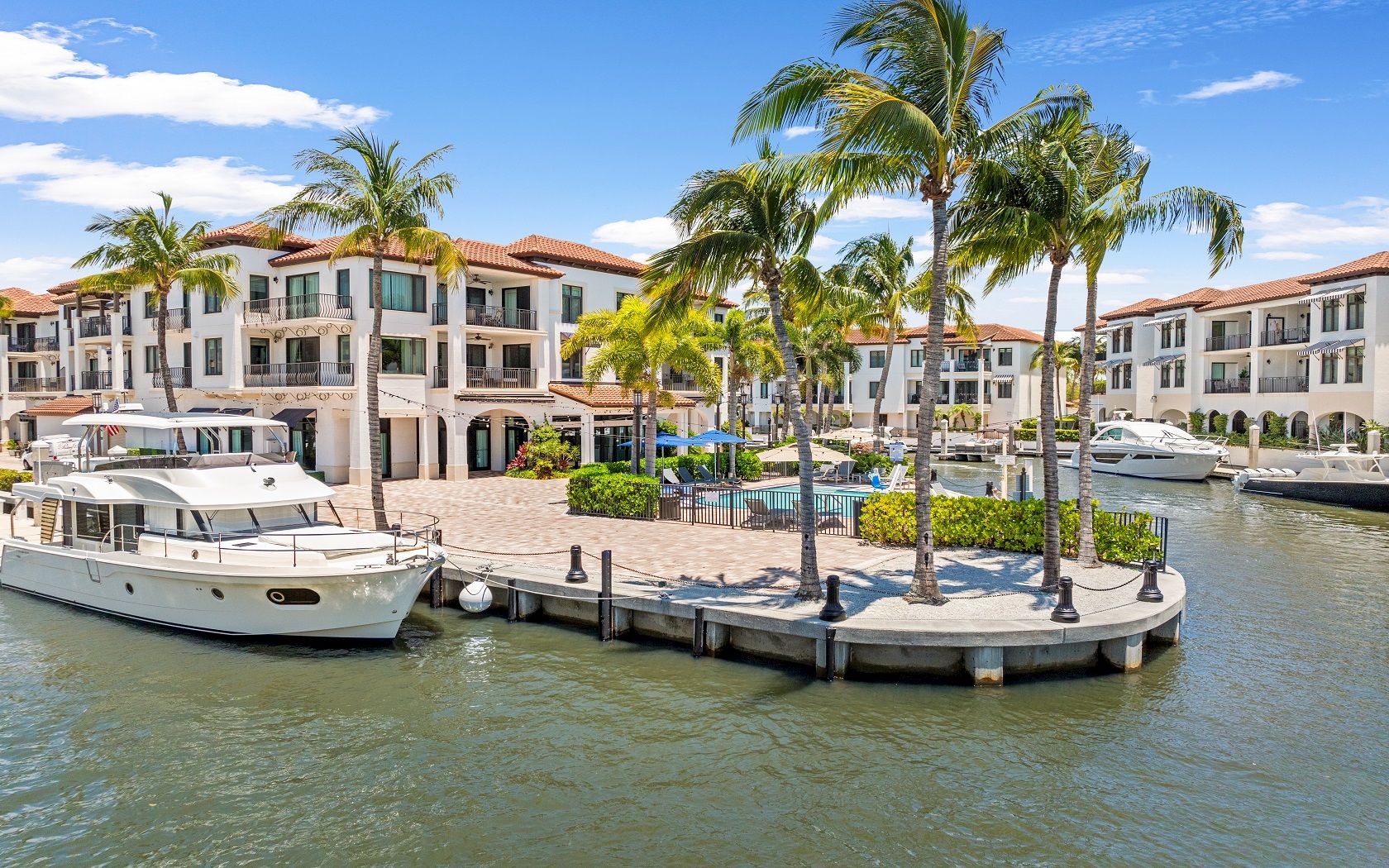 A marina with a small curved boardwalk, palm trees, boats docked, and a bright, sunny coastal apartment complex in the background.