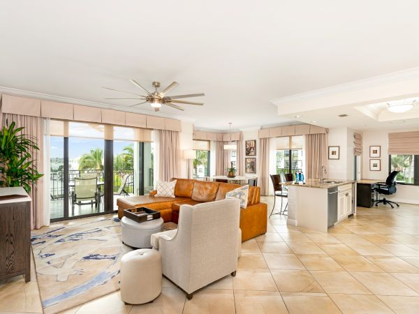 Bright open-plan living room with an orange sofa, tiled floor, large windows, ceiling fan, and a modern kitchen counter.