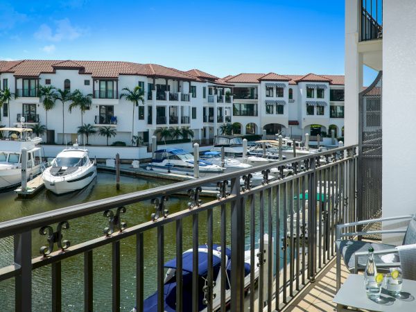 A sunny resort marina with white boats docked by a railing, palm-lined stucco buildings, and a balcony table in the foreground.