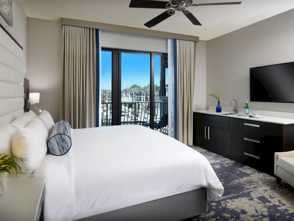 Modern hotel room with a king bed, balcony doors, curtains, flat-screen TV, and a ceiling fan overhead.