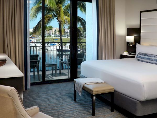 A bright hotel room with a king bed, small seating, and a balcony view of palm trees and a marina.