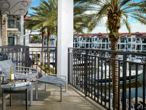 A sunny resort balcony with cushioned chairs and a bottle, overlooking palm trees, a courtyard, and a pool with boats.