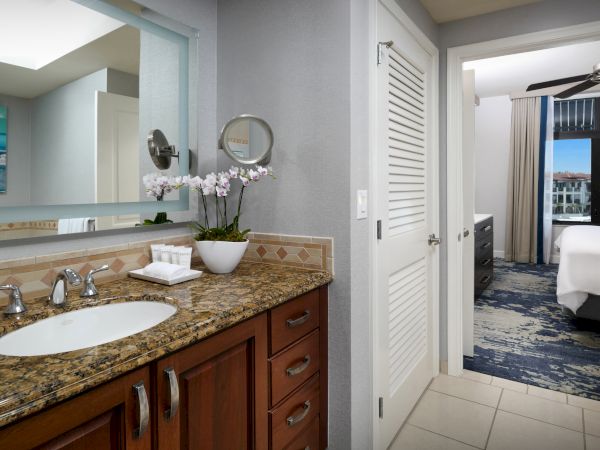Bright bathroom with a granite double vanity, orchid flowers, wall mirror, and an open doorway to a bedroom with curtains and a bed.