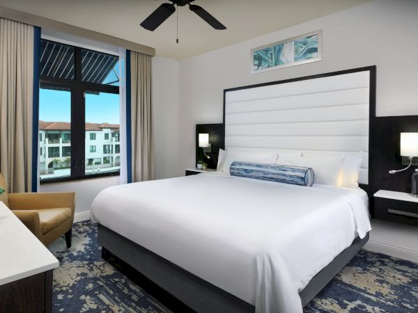 A modern hotel bedroom with a large bed, bedside lamps, wall art, and a window overlooking nearby buildings.