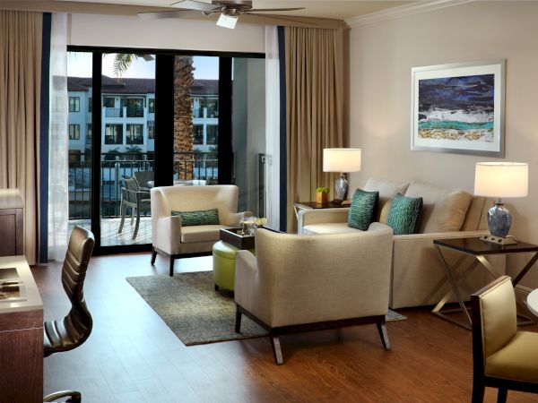 Sunlit living room with beige sofas, teal pillows, lamps, and floor-to-ceiling doors opening to a balcony courtyard view.