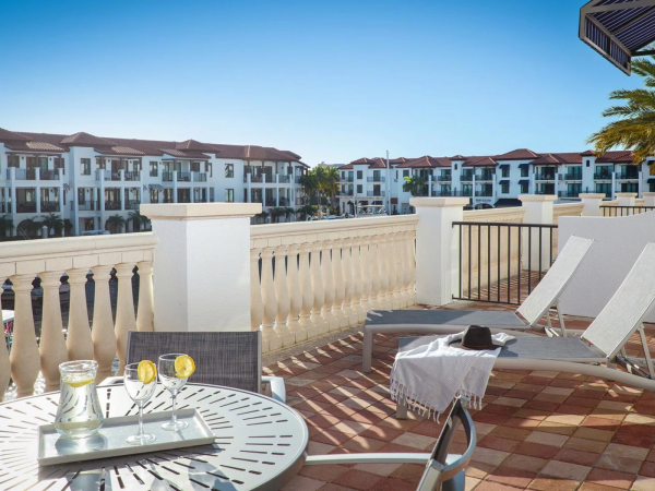 Sunny balcony at a resort with a patio table, drinks, and lounge chairs overlooking white apartment buildings and palm trees.