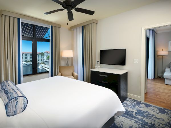 A bright hotel bedroom with a ceiling fan, TV on a dresser, curtains, and a view out the window.