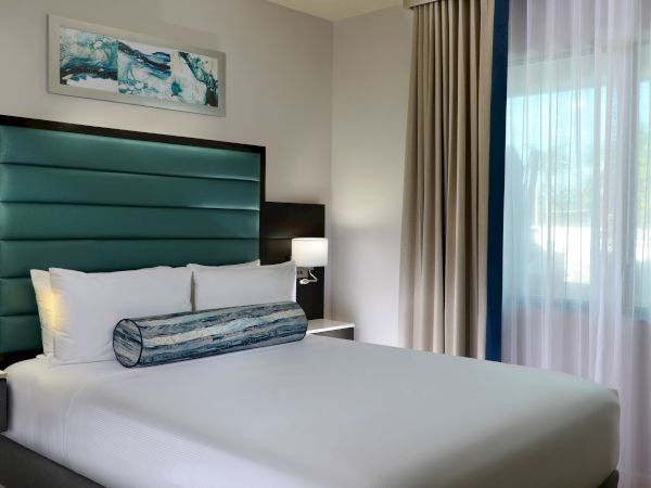 A modern bedroom with a teal upholstered bed, white bedding, side lamp, framed wall art, and beige curtains by a window.