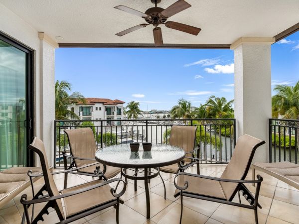 A sunny balcony with a round metal table, beige chairs, and a ceiling fan, overlooking palm trees and blue sky.