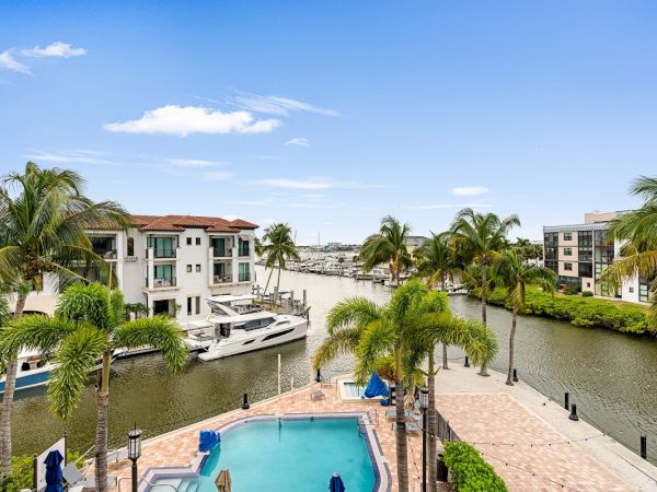 Palm-lined canal with boats, white buildings, and a turquoise swimming pool under a bright blue sky and clouds.