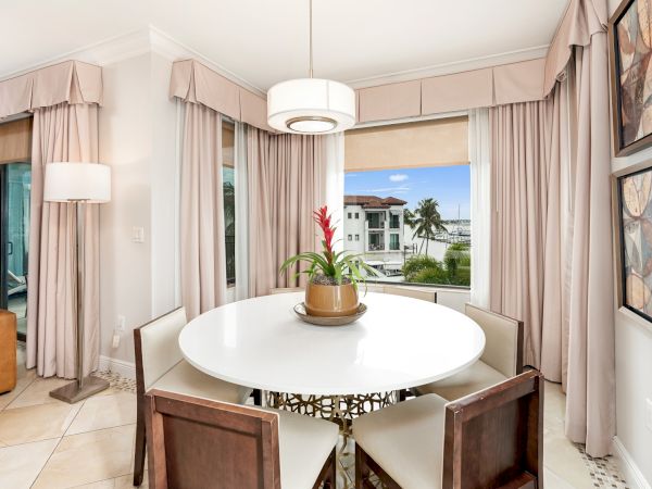 A bright dining area with round white table, pink curtains, ceiling light, and ocean-view windows.