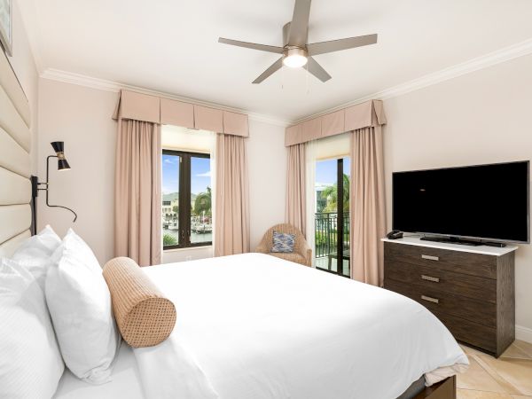 A bright bedroom with a ceiling fan, large bed, beige curtains, a TV on a dresser, and balcony doors to the view.