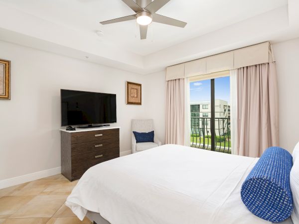 Modern bedroom with a ceiling fan, flat-screen TV on a dresser, and French doors to a balcony with beige curtains.