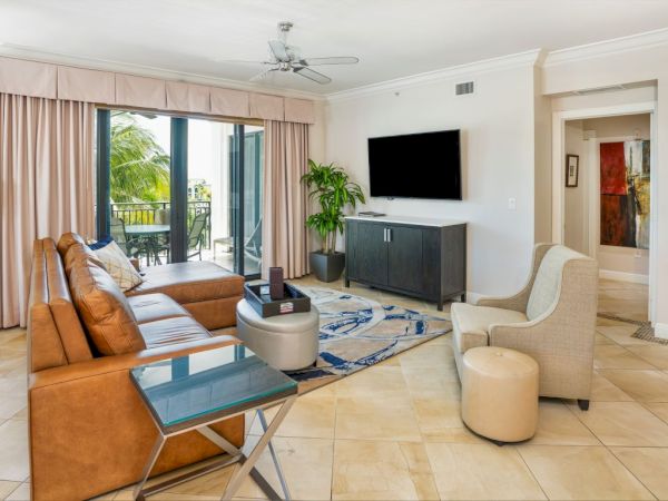 Bright living room with leather sofa, TV on a cabinet, rug, coffee table, and sliding doors to a balcony with palm views.