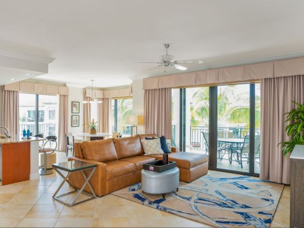 A bright open-plan living room and kitchen with a brown sectional sofa, TV, ceiling fan, and big sliding glass doors to a balcony.