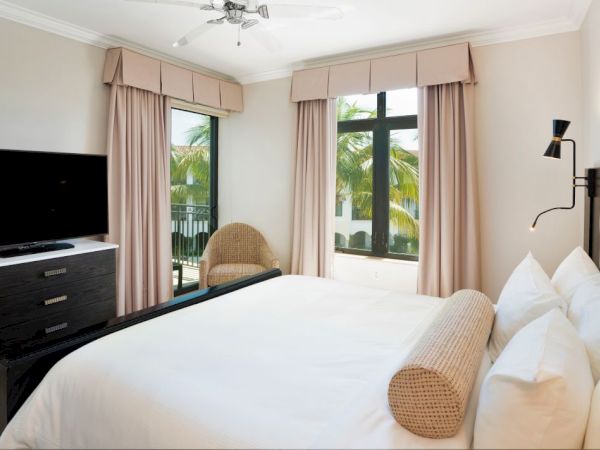 A bright modern hotel bedroom with a bed, TV dresser, and balcony doors framed by curtains and palm views.
