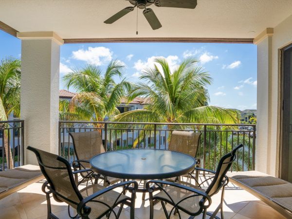 Sunny patio balcony with a round glass-top table, wicker chairs, and palm trees under a ceiling fan and clear blue sky.