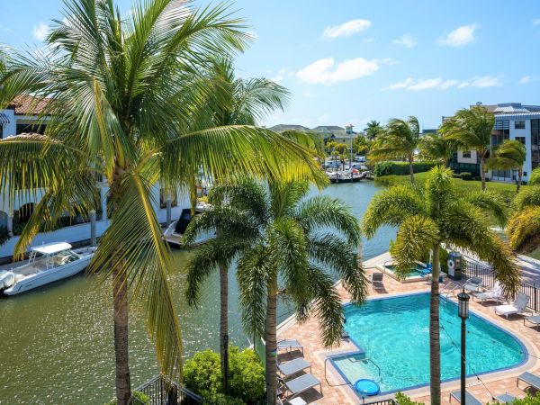 Tropical resort view with palm trees, a canal with boats, and a turquoise swimming pool beside white buildings under blue sky.