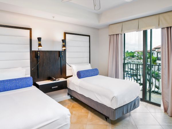 A bright hotel bedroom with two twin beds, bedside lamps, beige curtains, and a balcony view over palm-lined grounds.