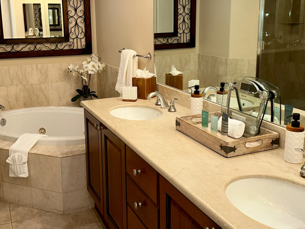 A luxurious bathroom with a double vanity, beige marble countertop, and a large mirror; a soaking tub, towels, and a tidy tray of amenities on the counter.