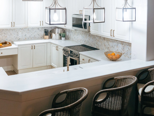 A bright, modern U-shaped kitchen with white cabinets, light backsplash, stainless appliances, and a breakfast bar with dark woven stools.