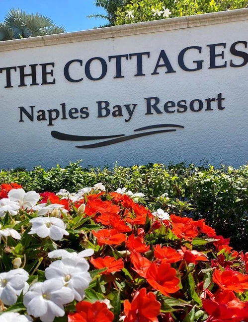 A sunny resort entrance sign reads &ldquo;THE COTTAGES, Naples Bay Resort,&rdquo; surrounded by colorful flowers and lush greenery.