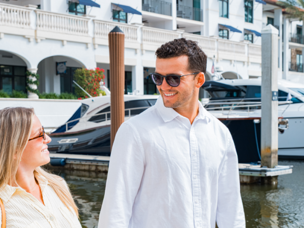 A smiling couple strolls on a marina dock by moored boats beside a bright waterfront hotel.