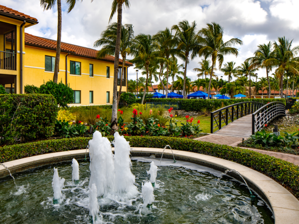 A sunny tropical resort courtyard with a stone fountain spraying water, palm trees, landscaped gardens, and yellow building walls.
