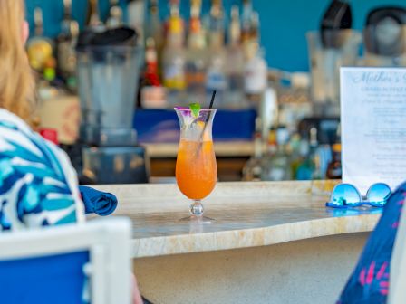 An orange cocktail in a glass with a garnish sits on a bar counter, with bottles and a menu blurred in the background.