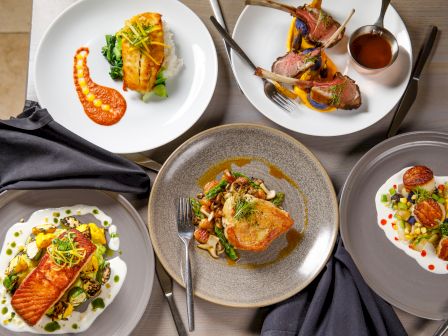 A gourmet spread of plated seafood and steak dishes with sauces, greens, and garnishes on three white plates.