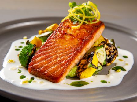 A plated grilled salmon fillet with crispy skin, saut&eacute;ed vegetables, microgreens, and a light herb oil drizzle.