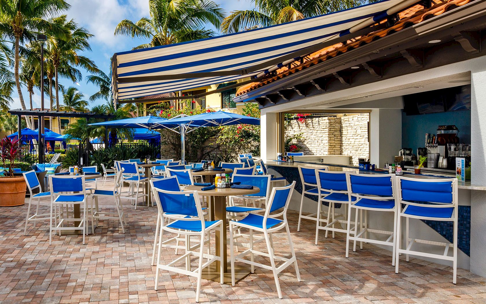 A sunny tropical bar scene with blue and white chairs, high-top tables, striped awnings, blue umbrellas, and palm trees in the background.