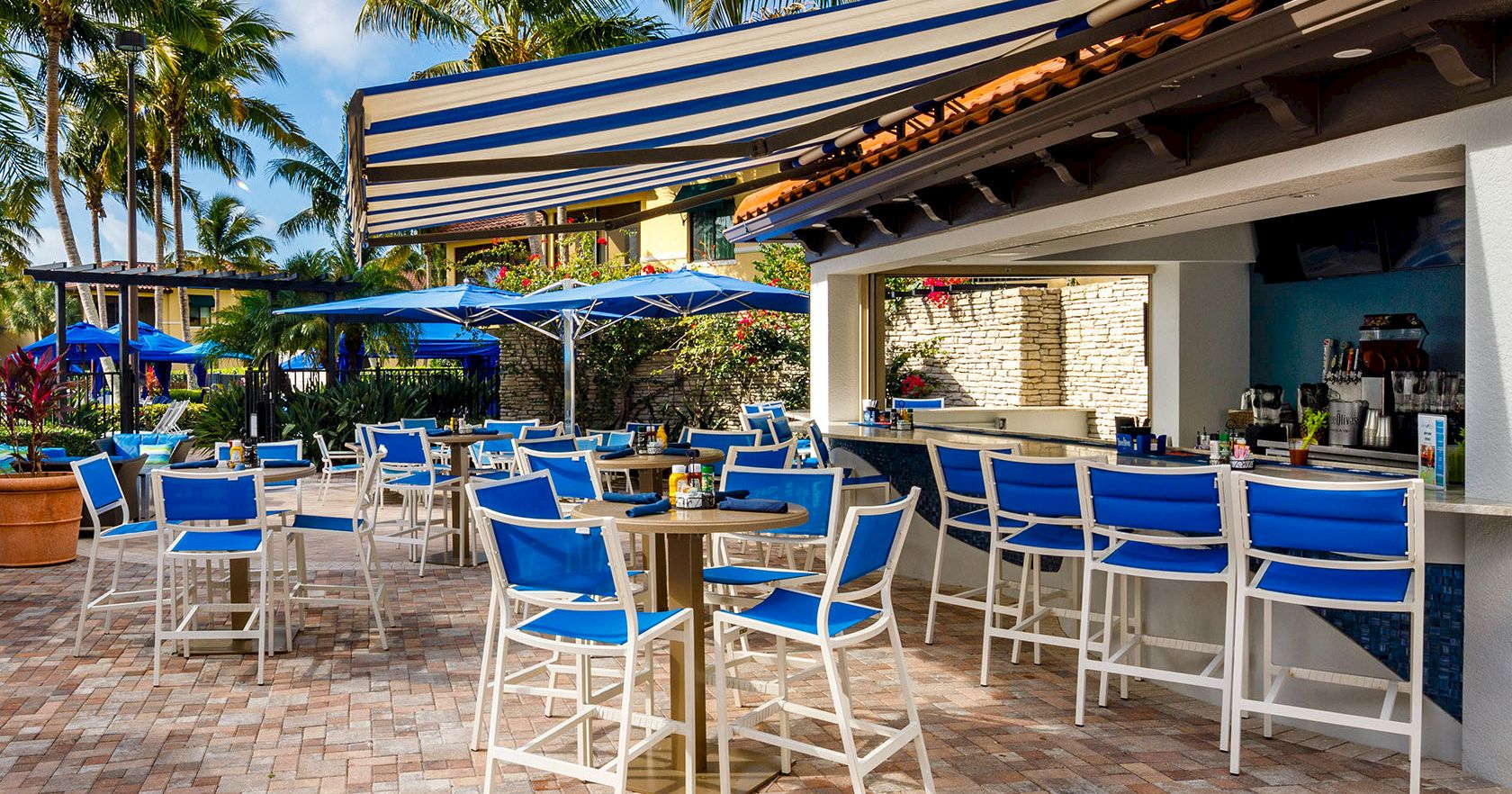 A sunny tropical bar scene with blue and white chairs, high-top tables, striped awnings, blue umbrellas, and palm trees in the background.