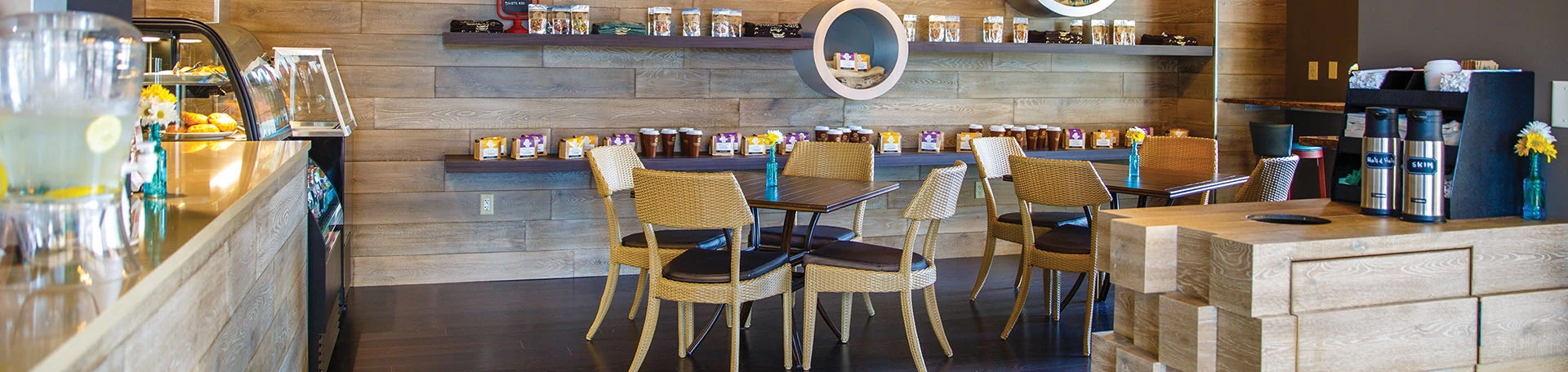 A cozy coffee shop interior with a pastry display case, small tables with chairs, and shelves of packaged treats and candles.