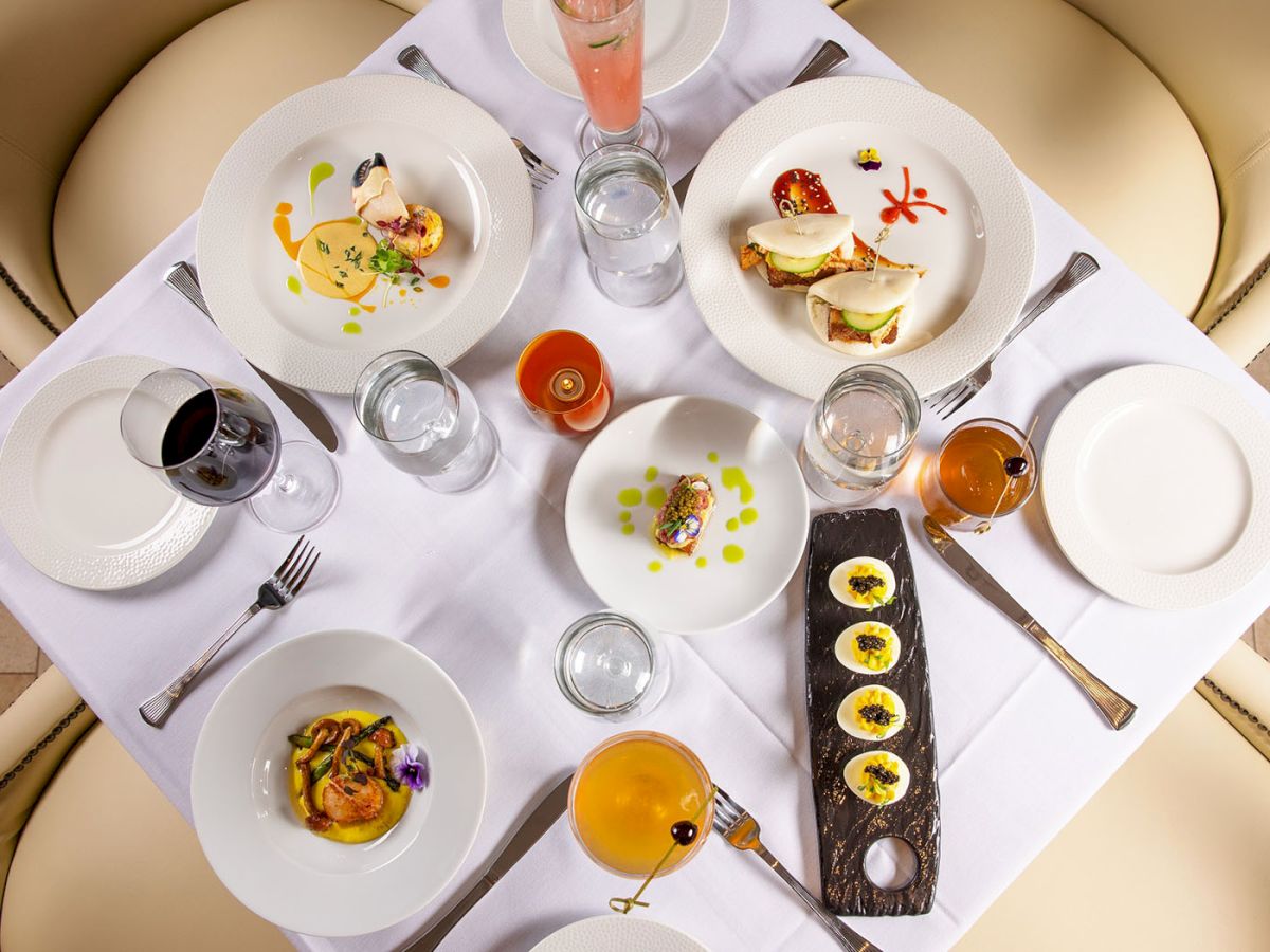 A gourmet restaurant table set with multiple plated dishes, drinks, and cutlery, arranged on a white tablecloth for a multi-course meal.