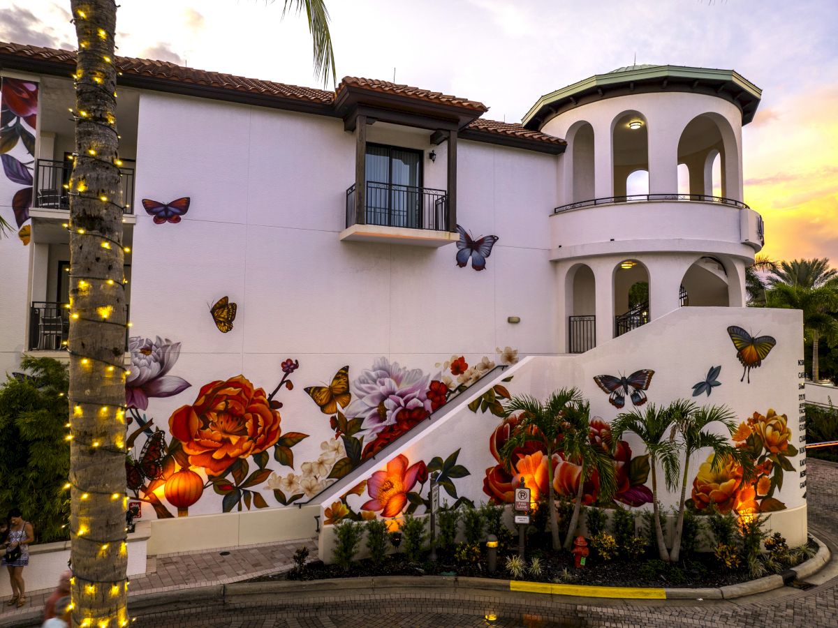 A white stucco building with balconies and arches, butterfly wall murals, palm trees, and warm string lights at sunset.