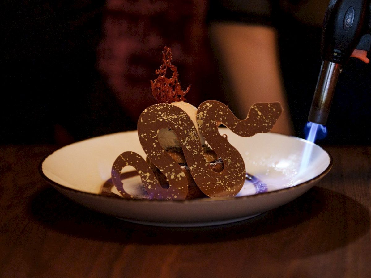 A chocolate &ldquo;100&rdquo; topped dessert with a lit sugar crown, while a blowtorch flame burns beside it on a plate.