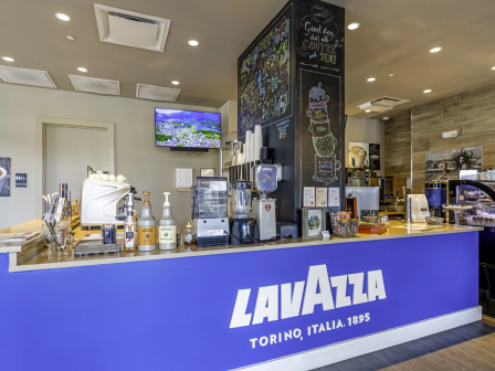 A Lavazza coffee shop counter with espresso machines, stacked cups, syrups, pastries, and menu displays in a bright caf&eacute;.