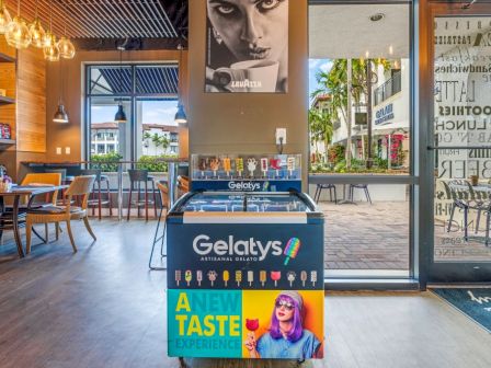 Gelato shop interior with Gelatys branding, tables and lamps, wall art, and a view of a sunny patio.