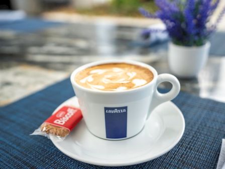 A Lavazza cappuccino sits on a patio table with a small pastry, while purple flowers blur in the background.