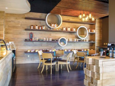 A cozy coffee shop interior with wooden shelves of snacks, round wall displays, hanging lights, and neatly set tables and chairs.