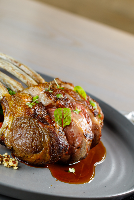 Plated roasted rack of lamb with green peas and asparagus, plus gravy, beside a filled wine glass.