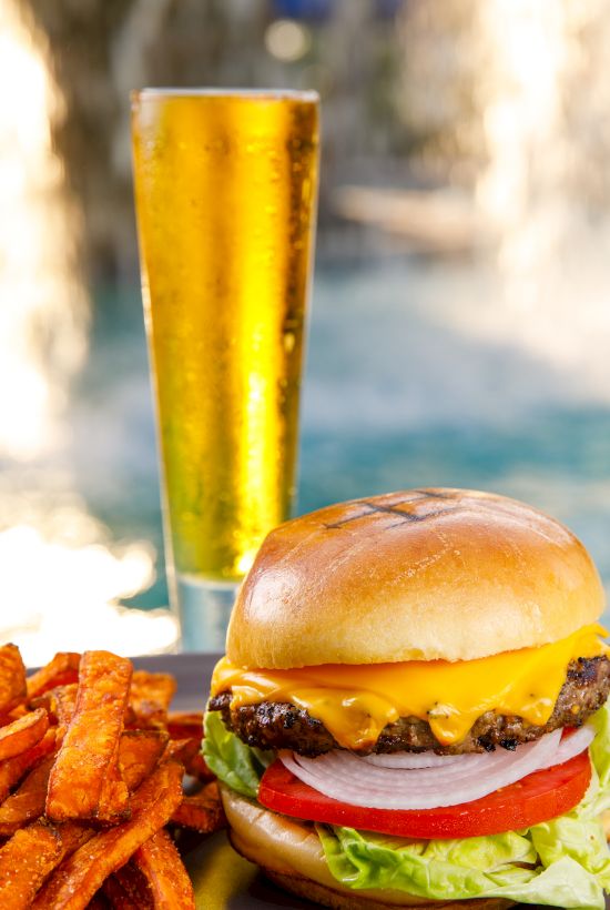 A juicy cheeseburger with lettuce and tomato, fries on the side, and a tall glass of golden beer in the background.